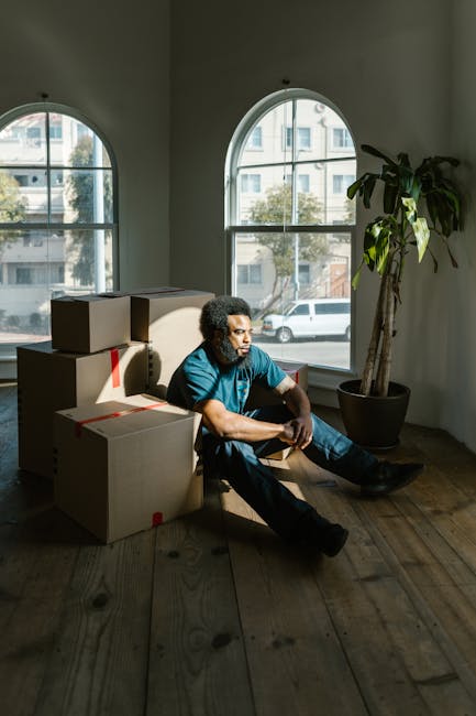 A man with curly hair, wearing a blue shirt and dark trousers, is sitting on a wooden floor inside a room with high, arched windows that let in natural light. He is surrounded by several cardboard moving boxes, some sealed with red packing tape and others open, indicating a packing or unpacking process as part of a house relocation. The boxes vary in size and are stacked in a casual manner behind and beside him. Next to him, there is a large potted plant with broad green leaves positioned near the window, adding a touch of interior decor. The room has plain white walls and a hardwood floor, providing a neutral environment typical of a residential relocation setting. Outside the windows, glimpses of urban buildings and parked cars are visible, suggesting the property is located in a city area such as Chelsea. The scene reflects the detail and professional approach offered by Man with Van Chelsea for home moving services, focusing on the logistics of furniture transport and packing within a residential environment.