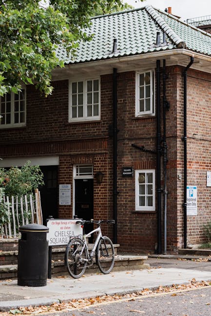 The exterior of a brick residential building on Chelsea Kings Road during a home relocation process. The building has a grey tiled roof, black drainpipes, and white-framed windows. A bicycle is parked against a black trash bin on the pavement outside, which is scattered with fallen leaves. A small, white roadside sign indicates parking restrictions for moving activities, with additional information posted on the building's wall. The scene suggests the involvement of Man with Van Chelsea in a furniture transport or packing and moving service, with a focus on local house removals in Chelsea. The environment is well-lit with natural daylight, and the setting shows a typical urban street scene suitable for loading and unloading furniture or boxes as part of home relocation logistics.