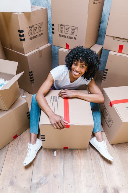 A young woman with curly black hair, wearing a white t-shirt, jeans, and white sneakers, is seated on a wooden floor surrounded by numerous brown cardboard boxes, some sealed with red packing tape and others open showing packing materials. She is smiling and holding a large box in her lap. The boxes vary in size and are stacked against a blue-painted wall, indicating an indoor setting typical of a home during a house removal or relocation process. In the background, additional boxes are visible, some placed on the floor, with a few partially opened, suggesting packing and loading activities. This scene reflects the typical environment during furniture transport and packing for a house move, relevant to services offered by Man with Van Chelsea, as seen on their website, manwithvanchelsea.co.uk, matching the context of the SW3 removals guide for best times for moving in Chelsea.