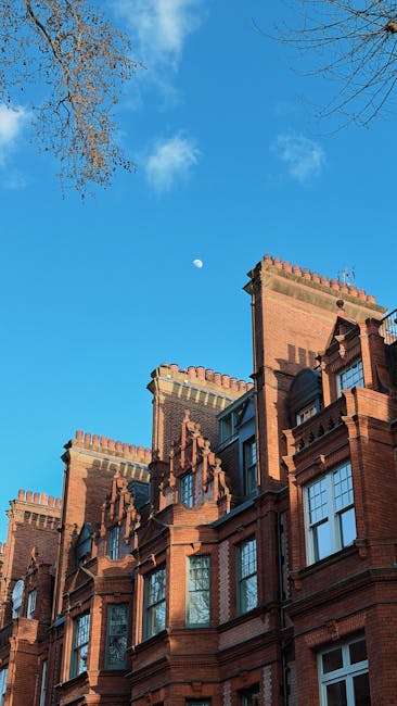 Photograph of a historic brick building in Chelsea under a bright blue sky with scattered clouds and a visible crescent moon. The building features multiple ornate chimneys, decorative brickwork, and large sash windows, some of which are open. Outside, several cardboard boxes, wrapped with plastic and secured with packing straps, are arranged on the sidewalk near a doorway, suggesting a home relocation or furniture transport process. A person from Man with Van Chelsea, a professional removals company, is seen lifting or carrying a large cardboard box towards a moving van parked nearby, which is partially visible with its open rear doors. The van is positioned on the pavement close to the entrance, with a trolley and moving blankets in the vicinity, indicating an active loading process during a house removal or furniture move in Chelsea. Natural lighting highlights the details of both the building and the packing materials involved in the packing and moving operation.