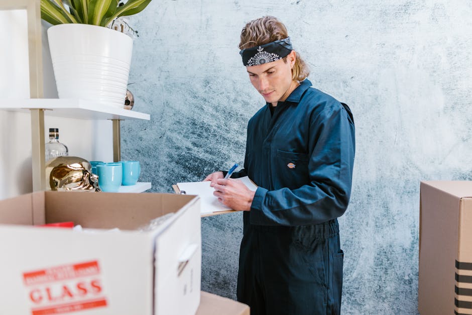 A young woman with curly black hair, wearing a white t-shirt, jeans, and white sneakers, is seated on a wooden floor surrounded by numerous brown cardboard boxes, some sealed with red packing tape and others open showing packing materials. She is smiling and holding a large box in her lap. The boxes vary in size and are stacked against a blue-painted wall, indicating an indoor setting typical of a home during a house removal or relocation process. In the background, additional boxes are visible, some placed on the floor, with a few partially opened, suggesting packing and loading activities. This scene reflects the typical environment during furniture transport and packing for a house move, relevant to services offered by Man with Van Chelsea, as seen on their website, manwithvanchelsea.co.uk, matching the context of the SW3 removals guide for best times for moving in Chelsea.